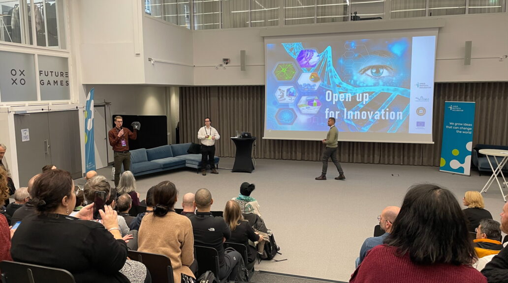 A filled room listening to a seminar. In the front it's a big screen.